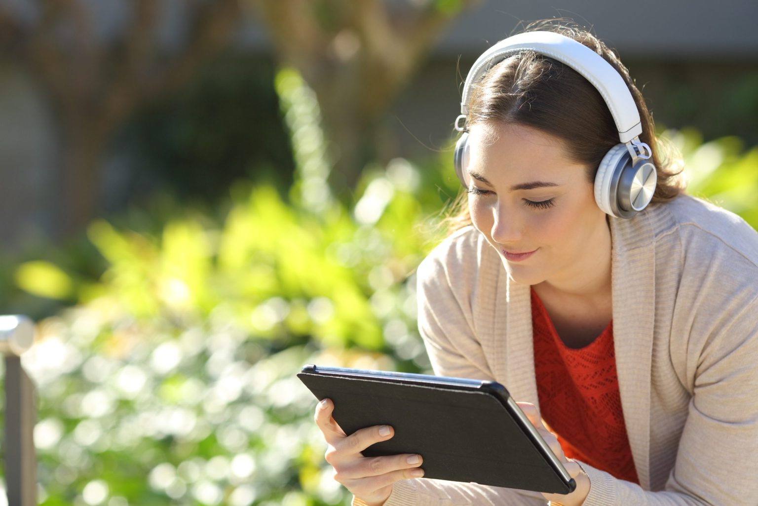 Woman sitting outside with headphones on, reading a tablet and smiling.
