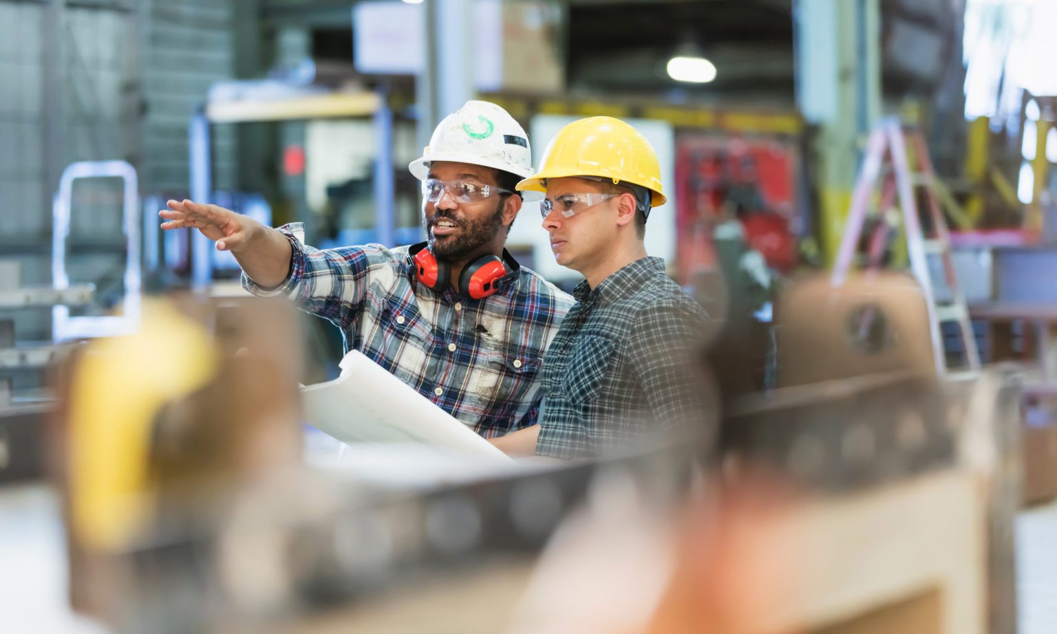 Two men in a warehouse wearing hard hats and safety glasses and one man is pointing.