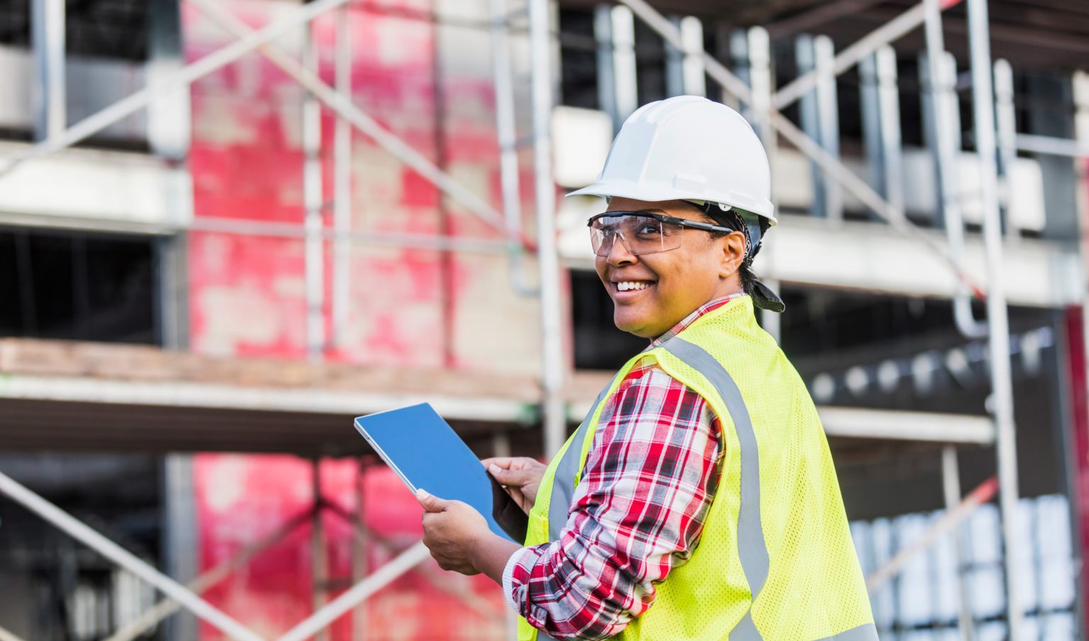 Woman construction worker at a construction site wearing a hard hat and safety glasses with a tablet and smiling.