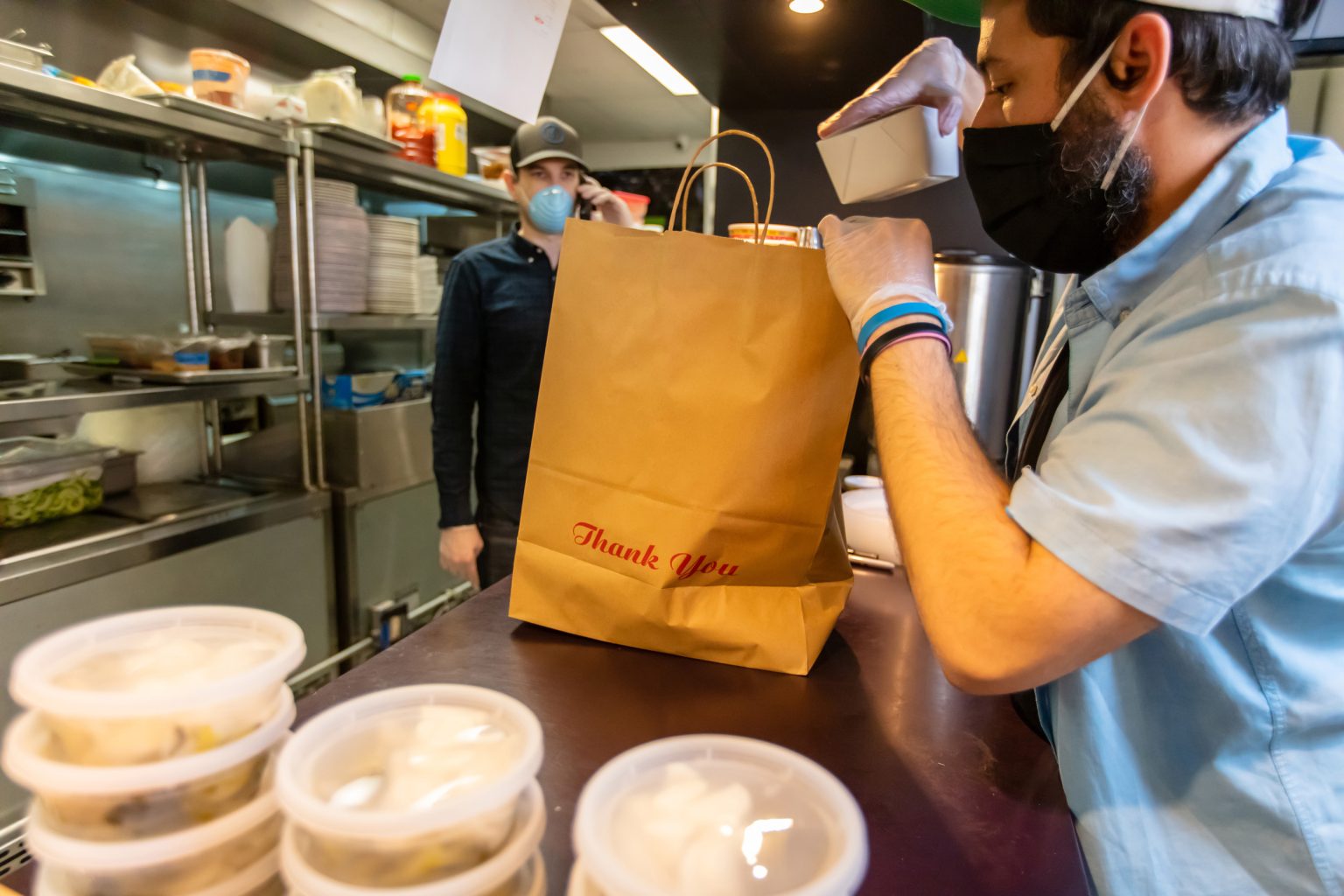 Two male food service workers in a restaurant kitchen both wearing masks. The one worker is filling up a paper bag with food.