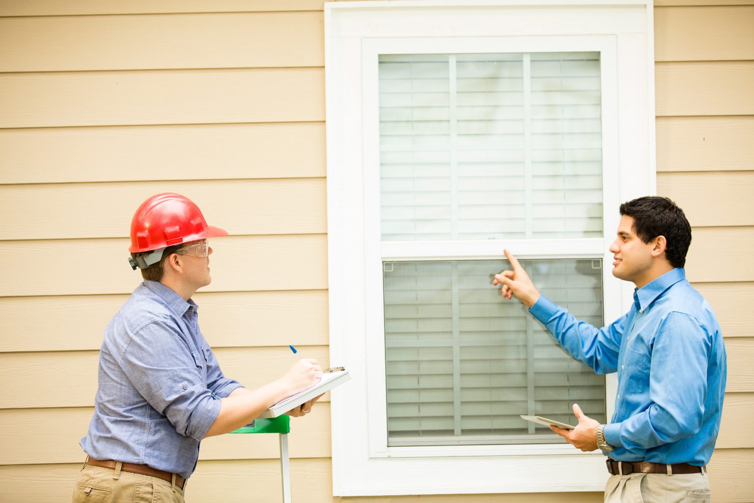 Two men outside of a window of a home and one is in a red hard hat and safety glasses and the other is pointing at the window.