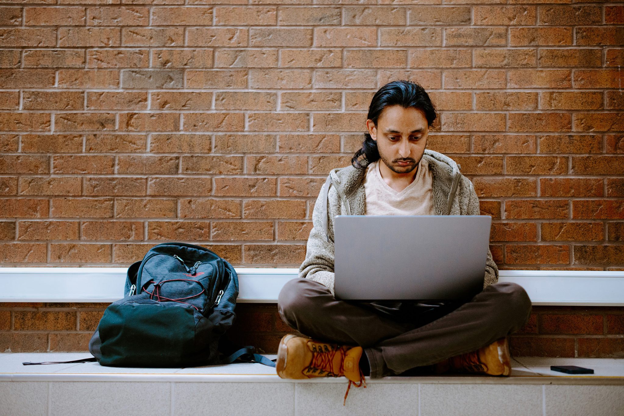 Male student sitting on the ground with a laptop and a bag beside.
